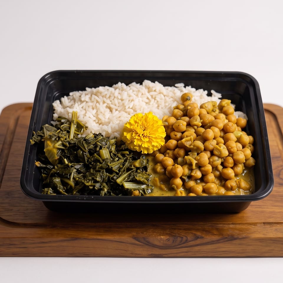 Meal tray with white rice, chickpea curry, sautéed leafy greens, and a yellow flower garnish on a wooden board.